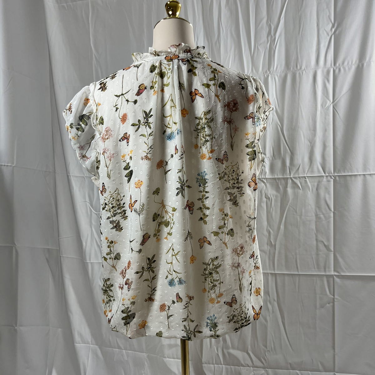 A white short-sleeve blouse with a colorful floral and butterfly print, displayed on a dress form mannequin against a wrinkled white backdrop.