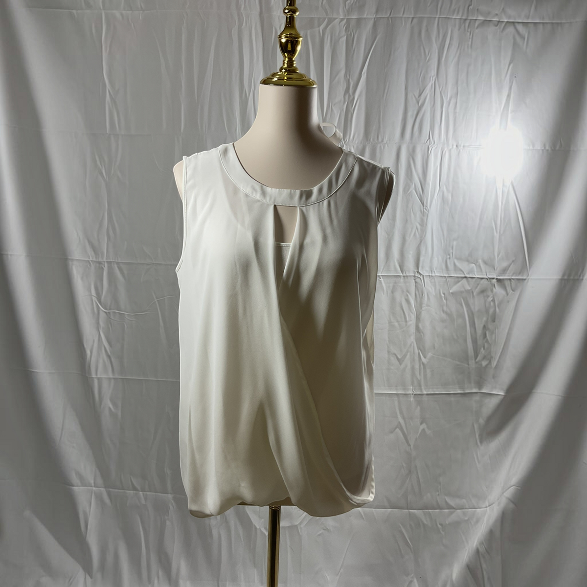 A sleeveless white blouse with a keyhole cutout at the neckline, displayed on a cream mannequin torso with a gold finial against a wrinkled white fabric backdrop.