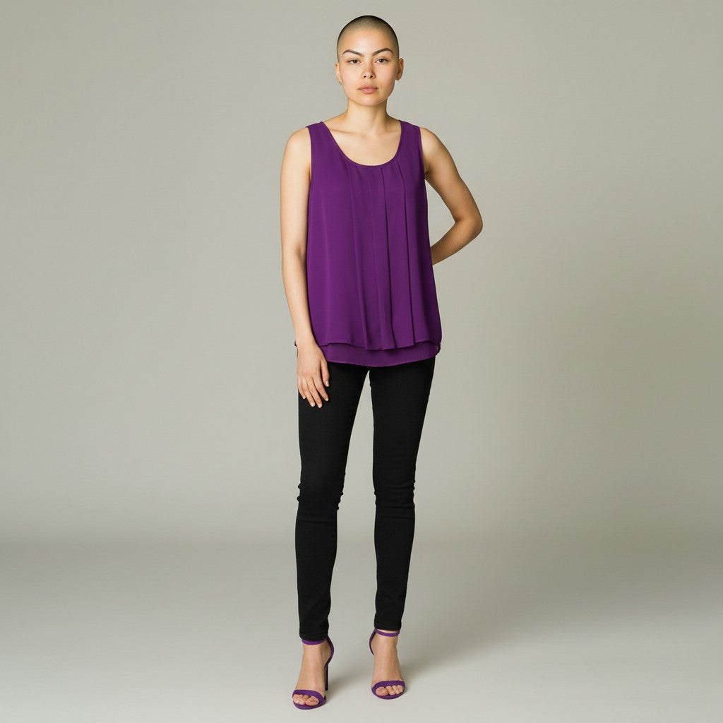 A model stands against a neutral light gray studio background wearing a sleeveless purple top with a draped, pleated design, black skinny pants, and purple high-heeled sandals.