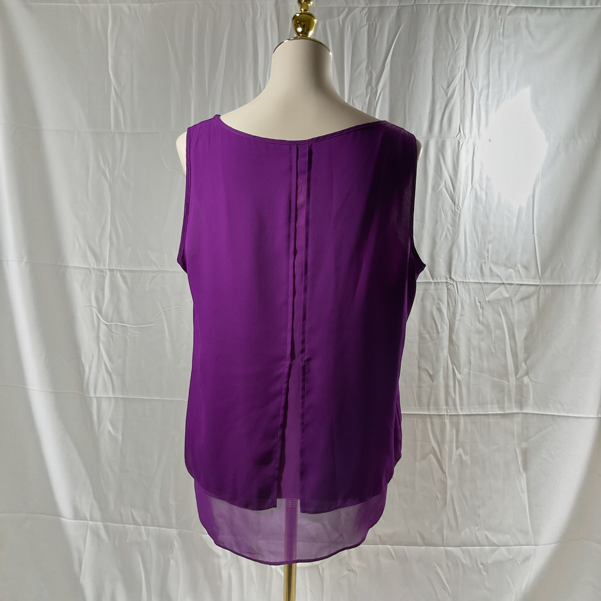 A sleeveless purple blouse displayed on a cream dress form mannequin, showing the back view with a sheer overlay hem, set against a wrinkled white fabric backdrop.