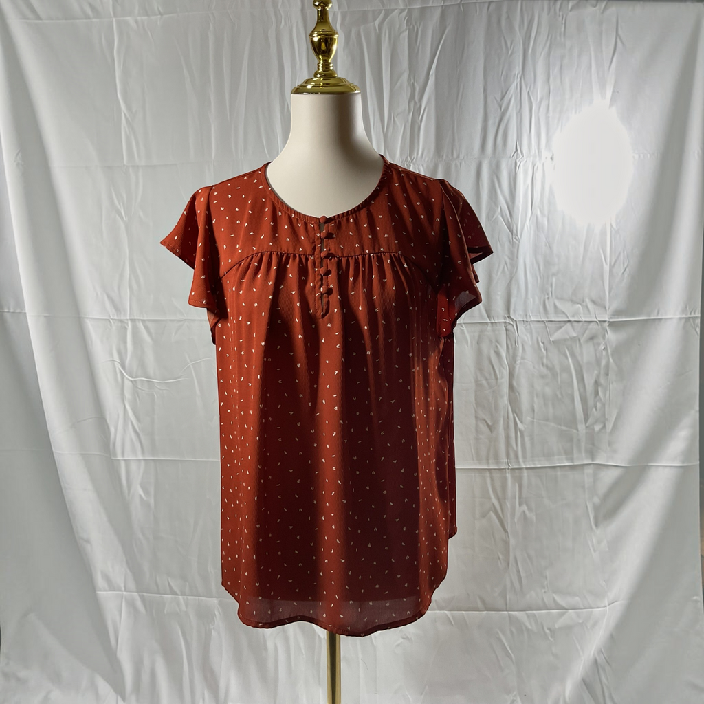 A rust-colored short-sleeve blouse with small white polka dots, displayed on a mannequin torso against a wrinkled white backdrop.