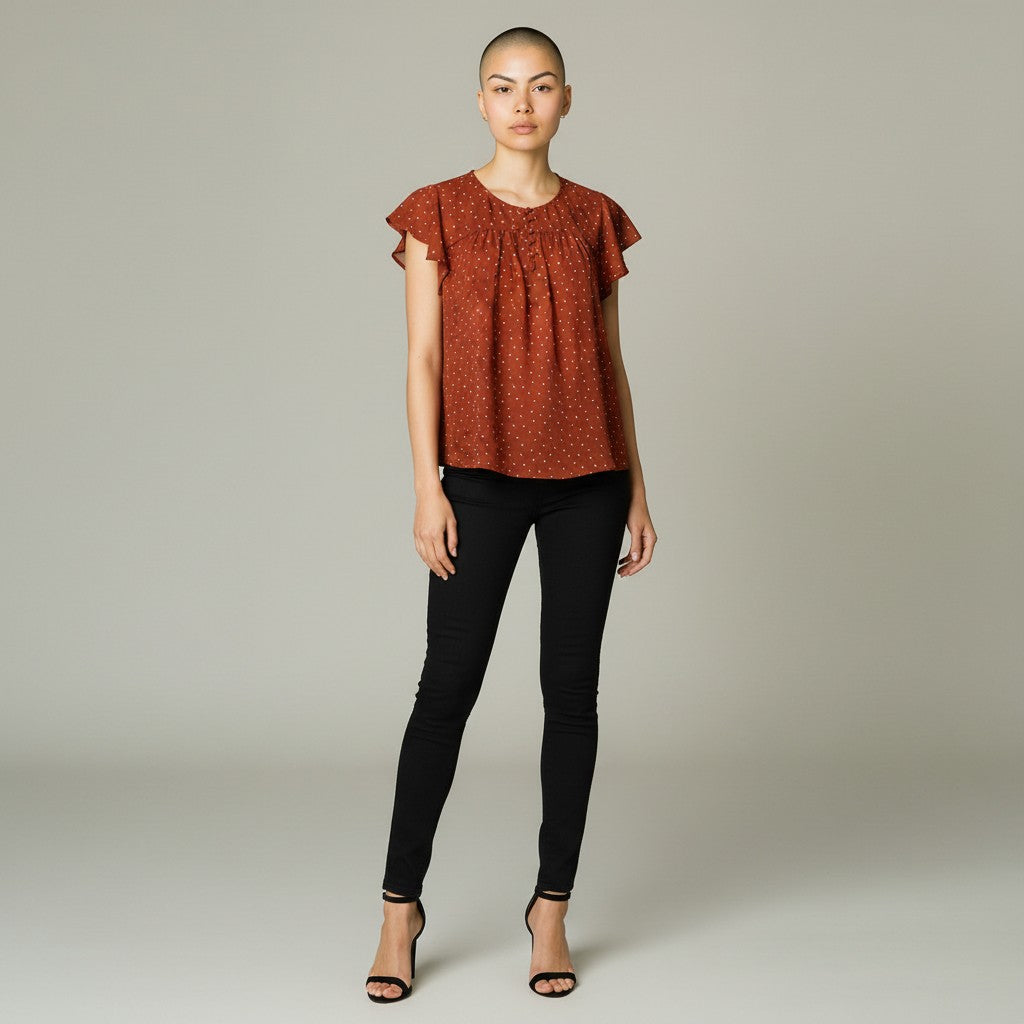 A woman modeling a rust-orange short-sleeve blouse with a small white pattern and ruffled shoulders, paired with black skinny pants and black strappy heels, against a neutral gray studio background.