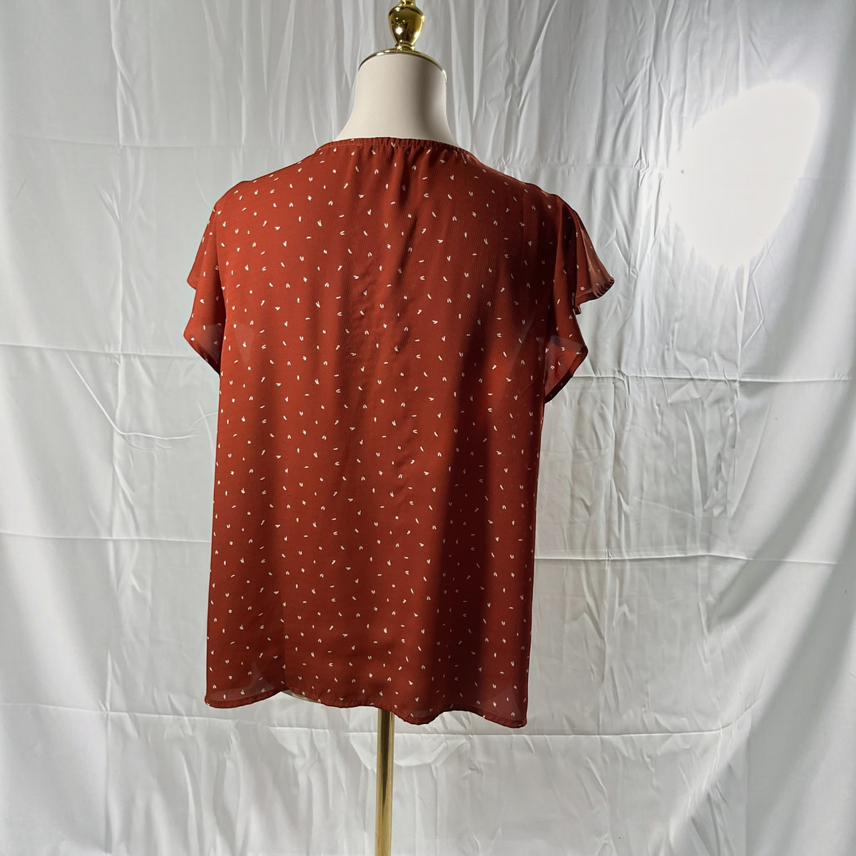 A rust-orange short-sleeve blouse with a white polka dot pattern, displayed on a dress form mannequin against a wrinkled white backdrop.