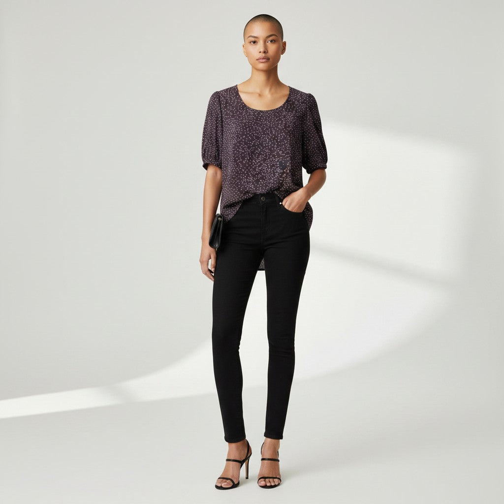 A woman in a dark gray shirt with white dots, black skinny jeans and black high heels on a studio gray background.