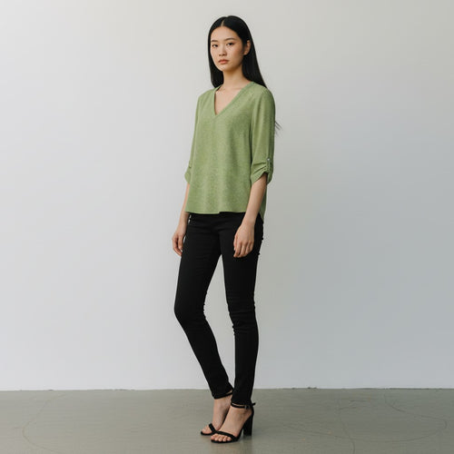 A woman modeling a light sage green knit V-neck top with rolled three-quarter sleeves, paired with black skinny jeans and black strappy heels, standing against a plain light gray wall.