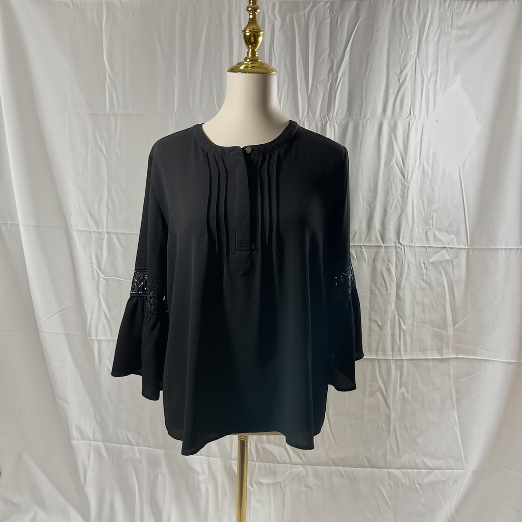 A black long-sleeve blouse with a round neckline, front pleats, and lace detailing on the cuffs, displayed on a mannequin torso against a white fabric backdrop.