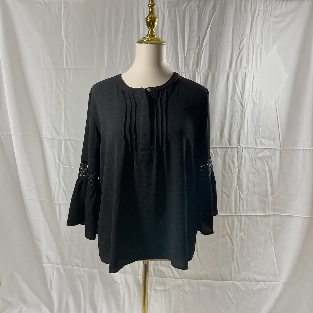 A black long-sleeve blouse with a round neckline, front pleats, and lace detailing on the cuffs, displayed on a mannequin torso against a white fabric backdrop.
