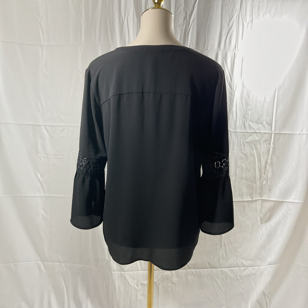 A black long-sleeve blouse displayed on a dress form from the back, featuring subtle lace detailing on the cuffs, set against a wrinkled white fabric backdrop.