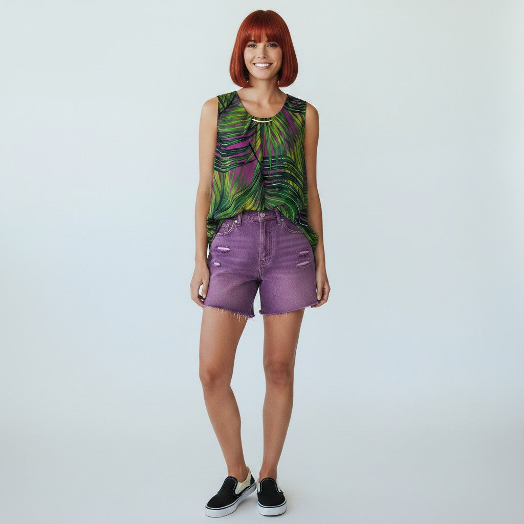 A woman with short red hair stands against a light gray studio background wearing a sleeveless top with a green, purple, and black abstract leaf-like print, purple denim shorts, and black slip-on sneakers.