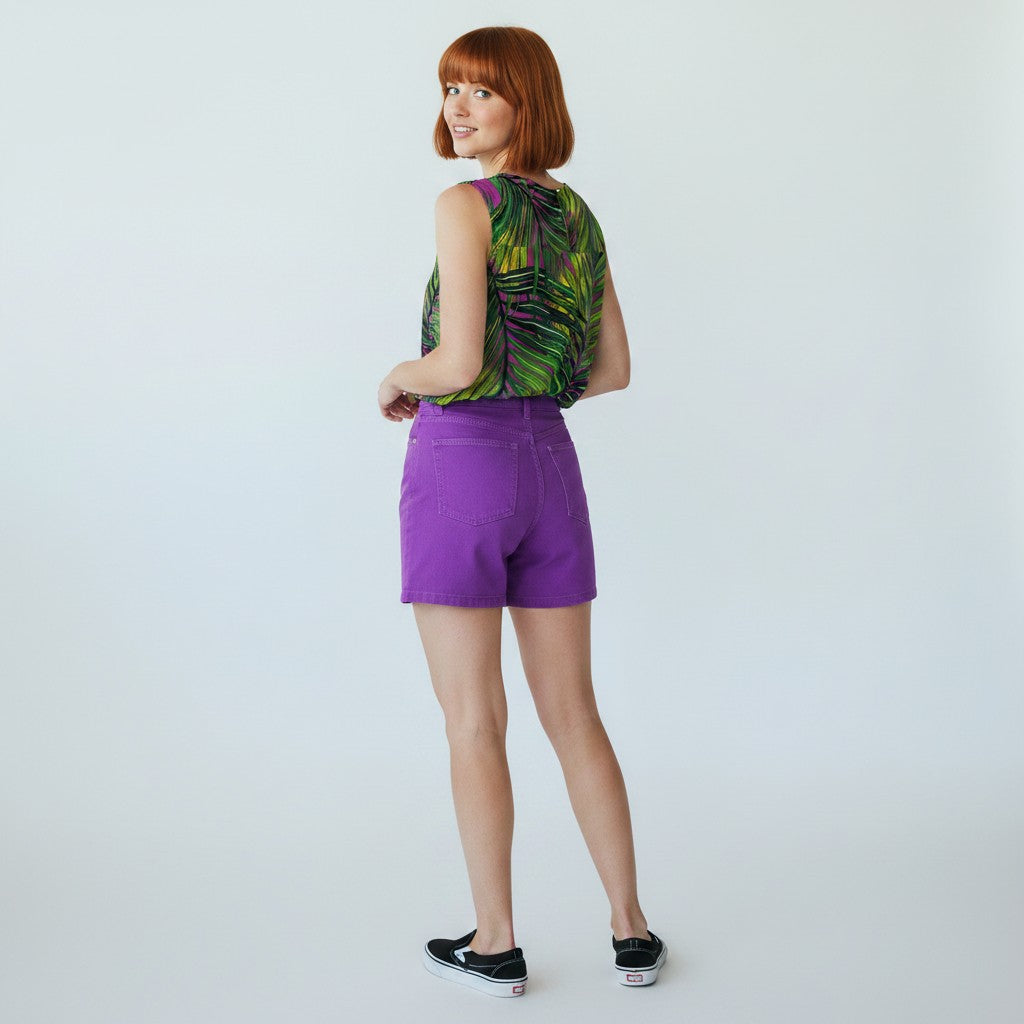 A woman with short red hair is shown from the back wearing a sleeveless green and black tropical leaf print top, bright purple shorts, and black slip-on sneakers against a light gray studio background.