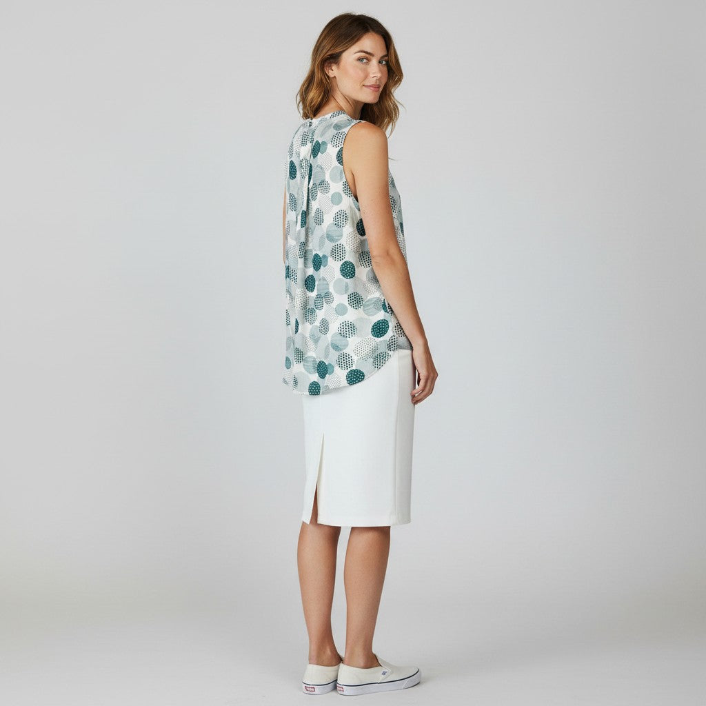 A woman stands in profile wearing a sleeveless top with a blue and green circular pattern on a white base, paired with a white knee-length skirt and white sneakers, against a light gray studio background.