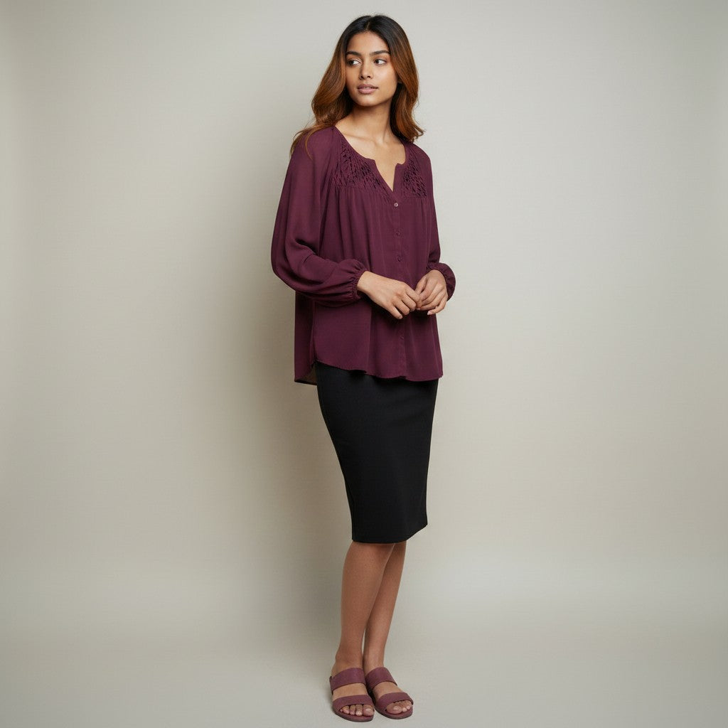 A woman modeling a long-sleeve burgundy blouse with lace detailing on the neckline, paired with a black knee-length skirt and mauve sandals, against a neutral studio backdrop.
