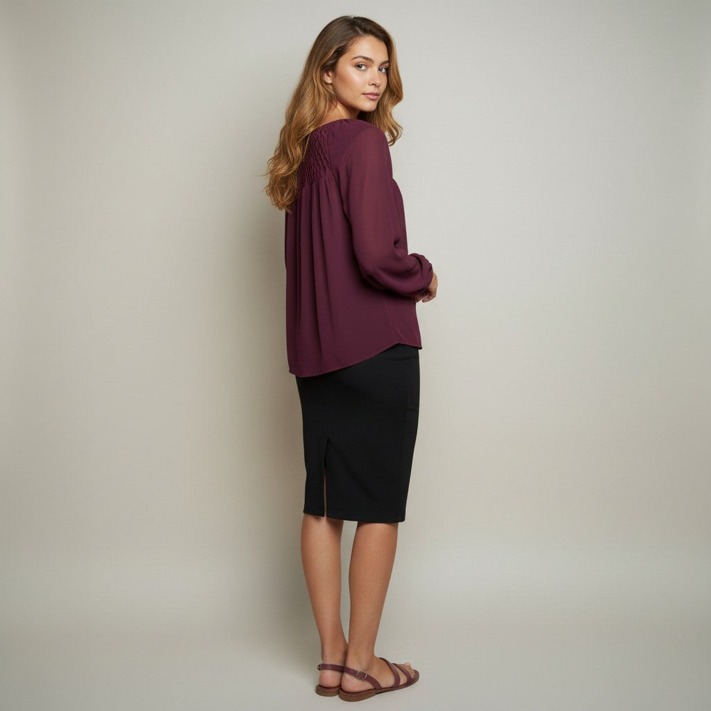 A woman stands in profile wearing a long-sleeve pleated top in deep burgundy and a black knee-length skirt, paired with brown strappy sandals, against a plain light gray studio background.