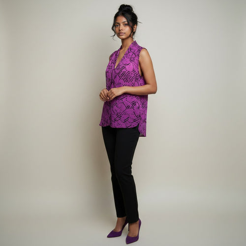 A woman modeling a sleeveless purple wrap-style top with a black abstract pattern, paired with black pants and purple heels, against a plain light beige studio background.