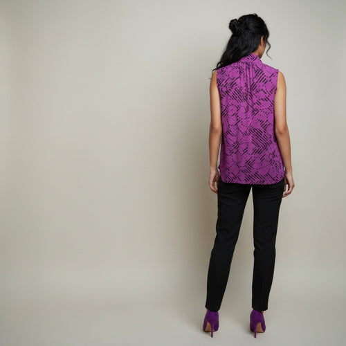 A woman shown from the back wearing a sleeveless high-neck top with a purple and black abstract pattern, paired with black pants and purple heels, against a plain light beige studio background.