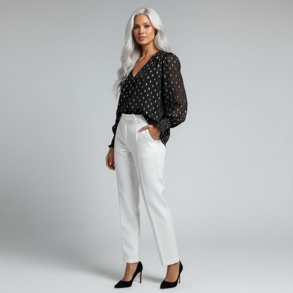 A woman with long silver hair models a black sheer long-sleeve blouse with a white polka-dot pattern, paired with white trousers and black heels, against a light gray studio background.