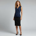 A woman modeling a sleeveless navy blue wrap-style top paired with a black knee-length skirt and black heels, standing against a light gray studio background.