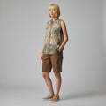 A woman modeling a sleeveless button-front shirt with an abstract animal-like print in beige, brown, and black, paired with brown shorts and tan slip-on shoes, against a light gray studio background.