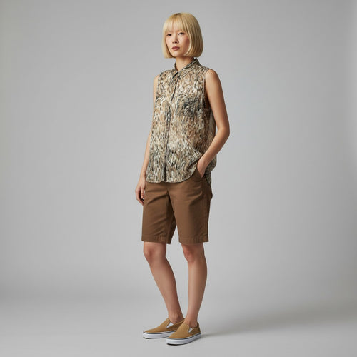 A woman modeling a sleeveless button-front shirt with an abstract animal-like print in beige, brown, and black, paired with brown shorts and tan slip-on shoes, against a light gray studio background.