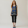 A woman with red hair stands against a light gray studio background wearing a sleeveless V-neck top with a blue, black, and silver ornate pattern, paired with black cropped pants and blue heels.