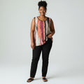 A woman modeling a sleeveless top with an abstract vertical stripe pattern in pink, red, orange, and cream, paired with black pants and sandals, against a light gray studio background.