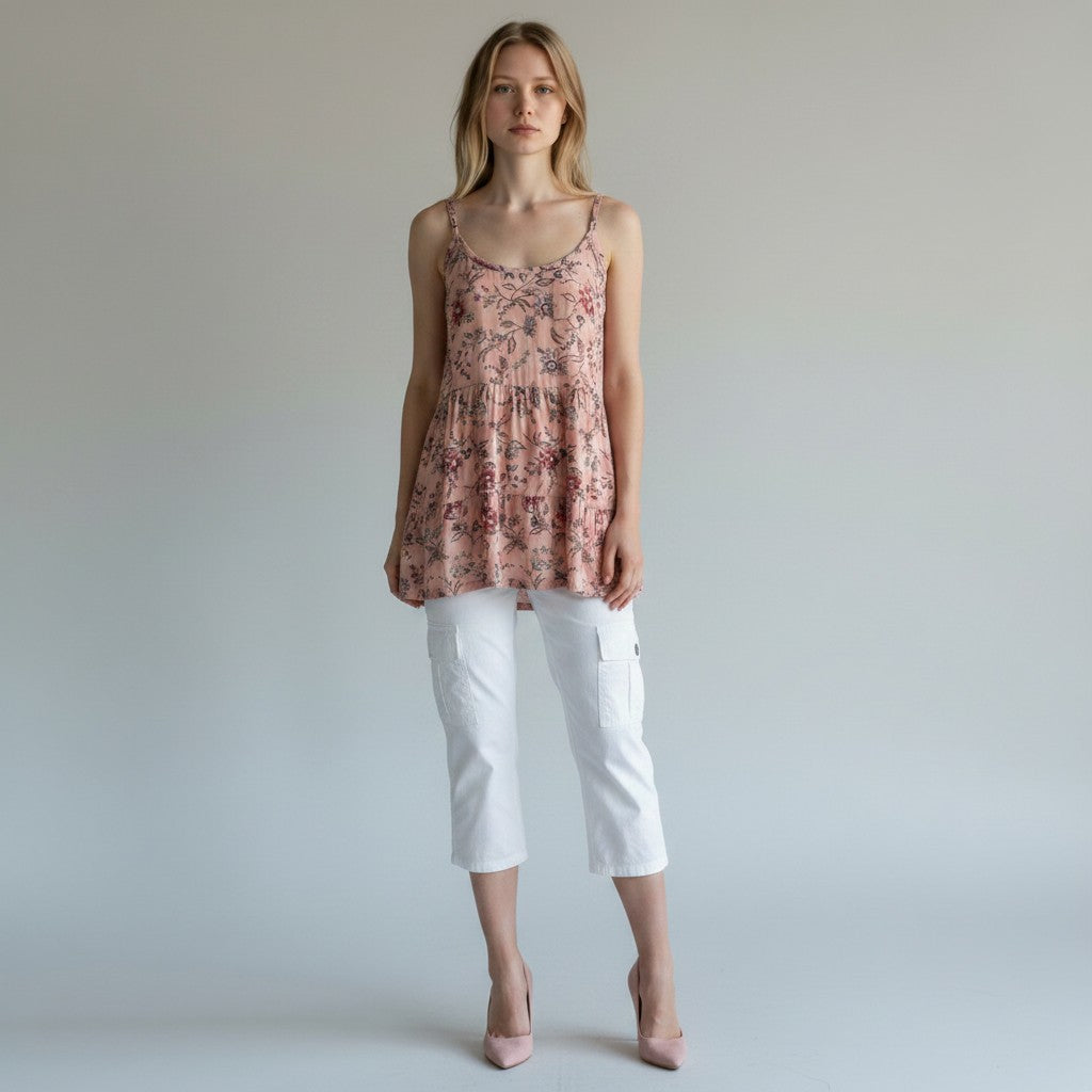 A woman modeling a sleeveless floral peplum top in dusty pink and red tones, paired with white cropped pants and nude heels, against a plain light gray studio background.