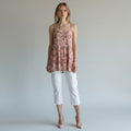 A woman modeling a sleeveless floral peplum top in dusty pink and red tones, paired with white cropped pants and nude heels, against a plain light gray studio background.