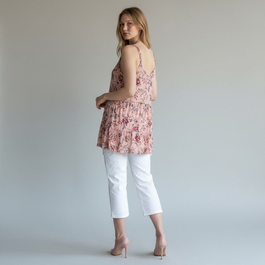 A woman stands in profile wearing a light pink floral camisole top with white cropped pants and nude heels against a plain light gray studio background.