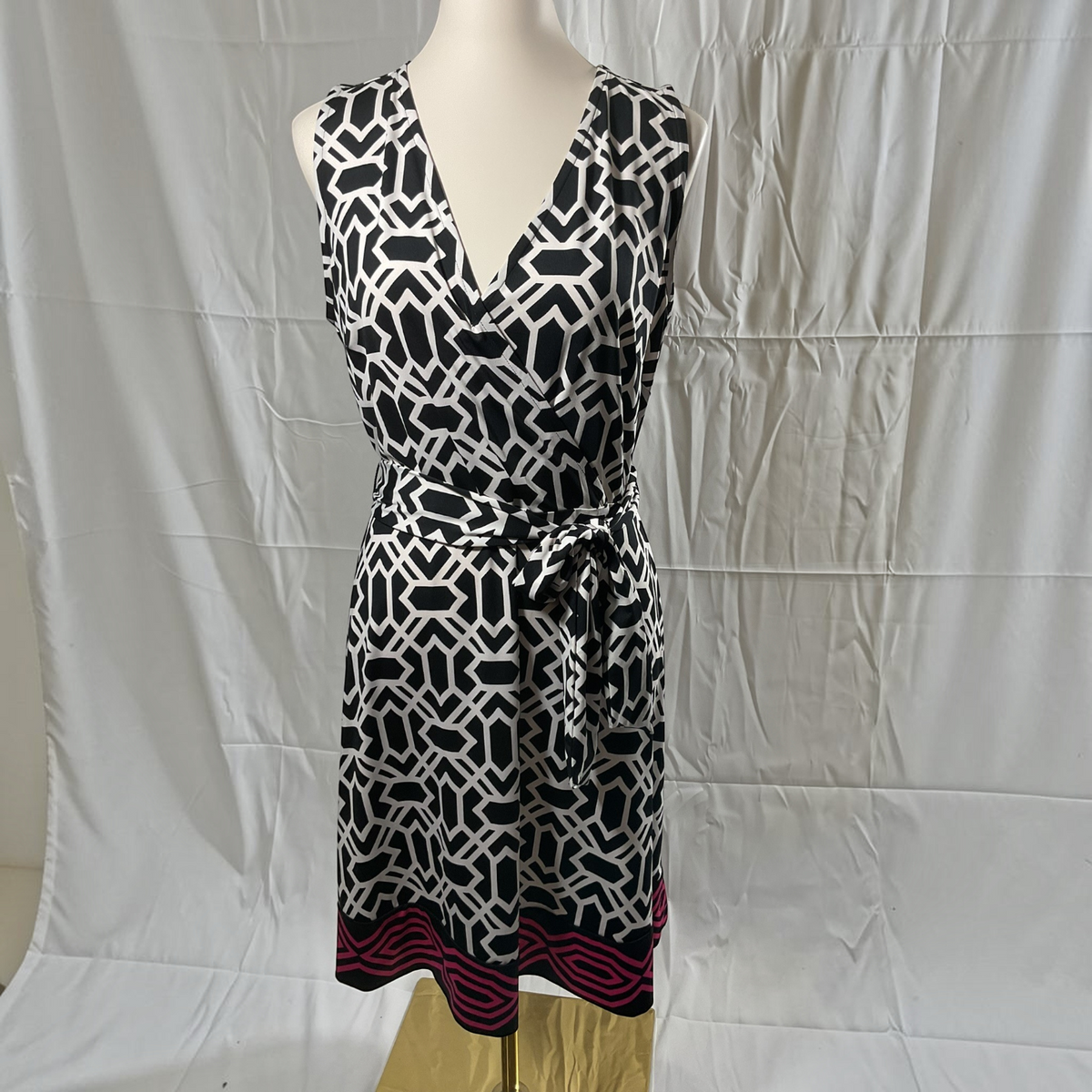 A sleeveless V-neck dress with a black and white geometric pattern and a magenta striped hem, displayed on a mannequin against a white fabric backdrop.
