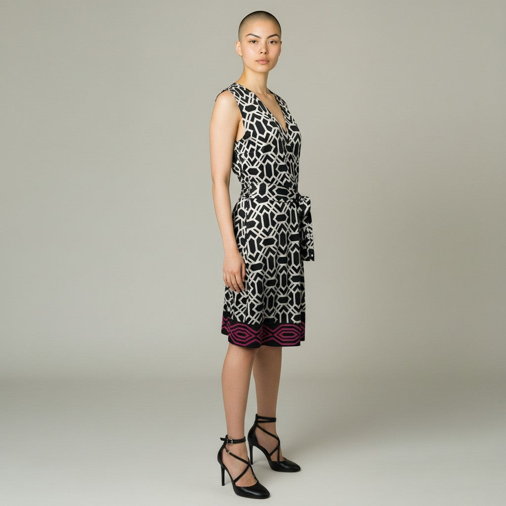 A model wearing a sleeveless wrap-style dress with a black and white geometric pattern and a magenta border at the hem, paired with black strappy heels, against a neutral gray background.