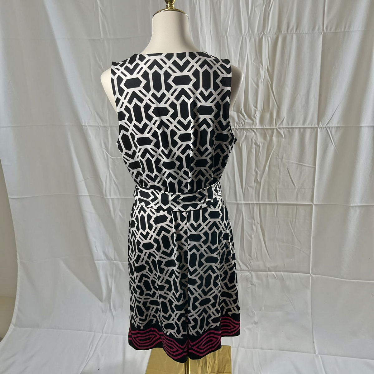 A sleeveless black and white geometric print dress displayed on a mannequin, featuring a matching waist belt and a pink striped hem, set against a white fabric backdrop.
