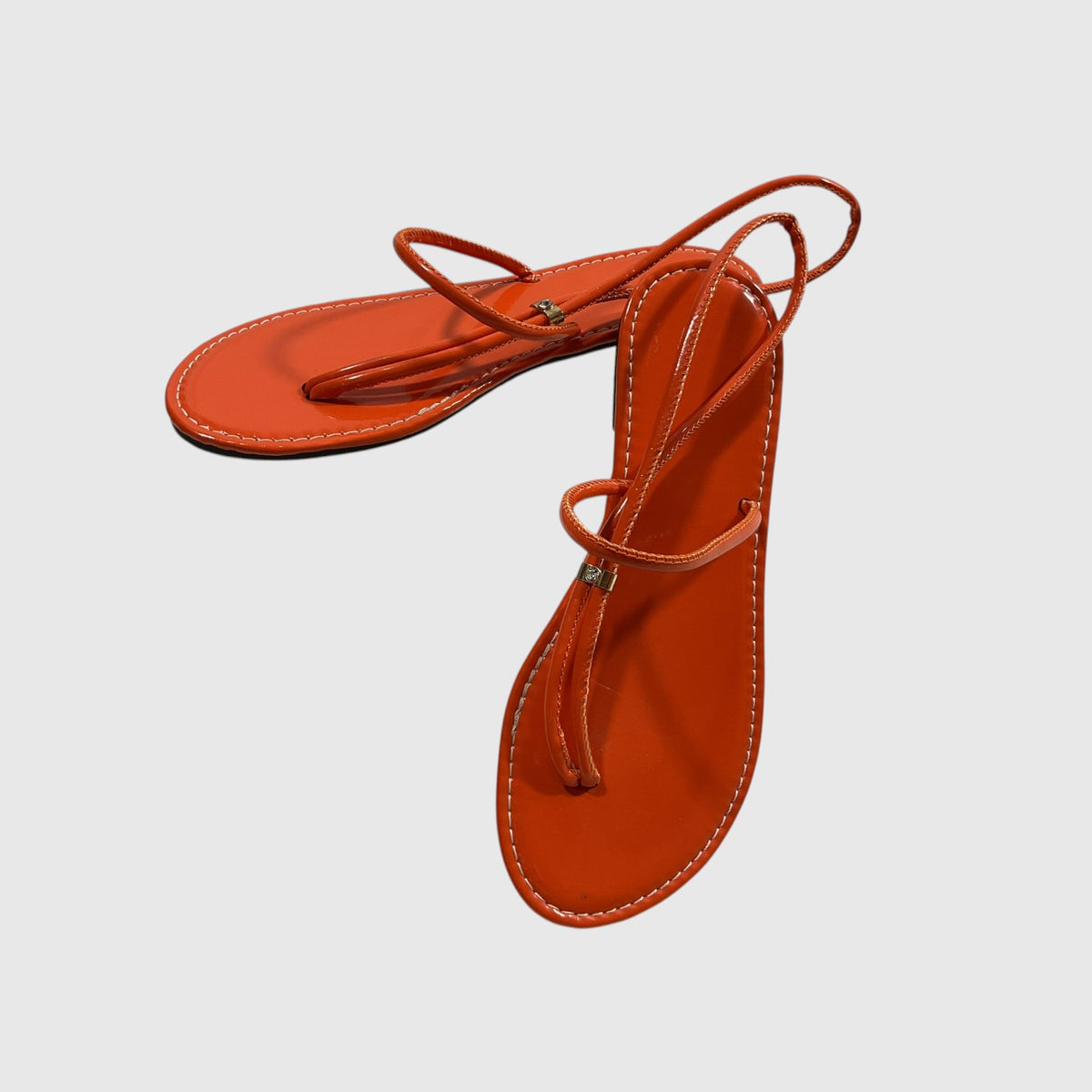 A pair of women's minimalist flat sandals in a vibrant burnt orange color, featuring thin wrap-around straps and small gold-tone decorative studs, shown on a light gray background.