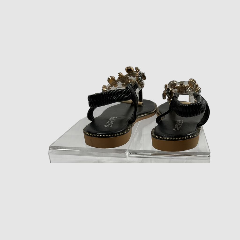 A pair of black flat sandals with textured straps and gold-tone floral embellishments, displayed on a clear acrylic block against a light gray background.