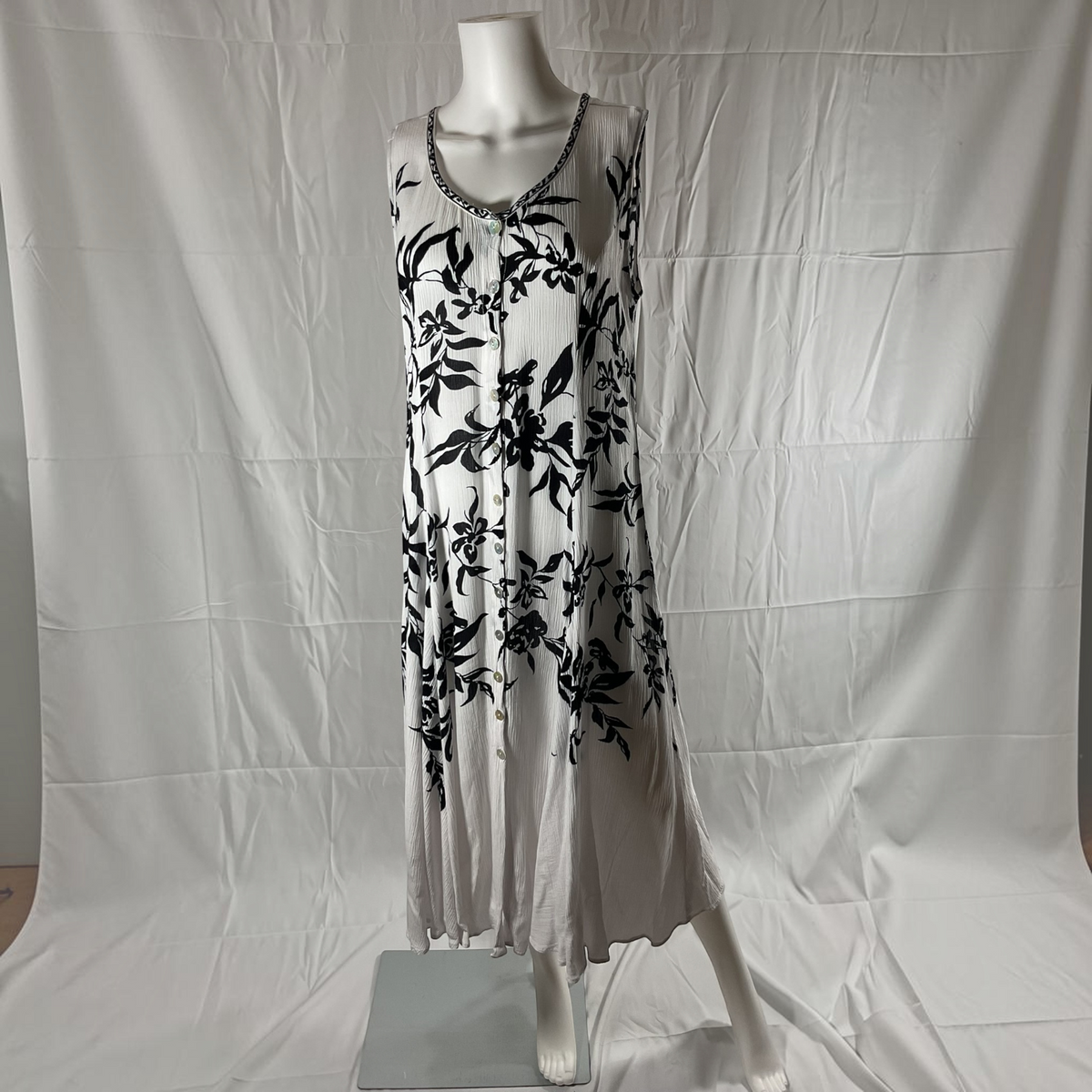 A sleeveless white midi dress with a black floral print and a beaded neckline, displayed on a white mannequin against a wrinkled white backdrop.
