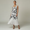 A woman modeling a sleeveless white midi dress with a black floral print, standing against a neutral light gray studio background.