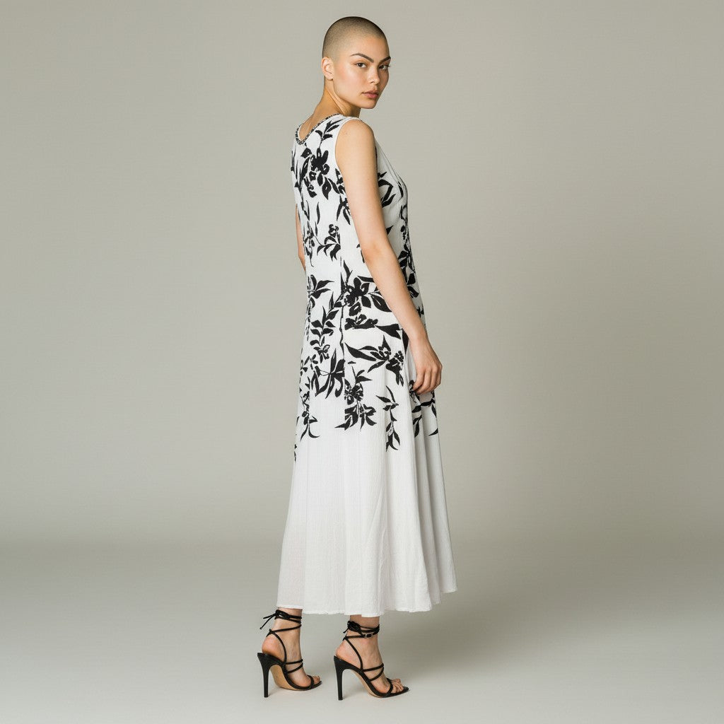 A woman modeling a sleeveless white maxi dress with a black floral print, shown from a three-quarters back view against a neutral light gray studio background.