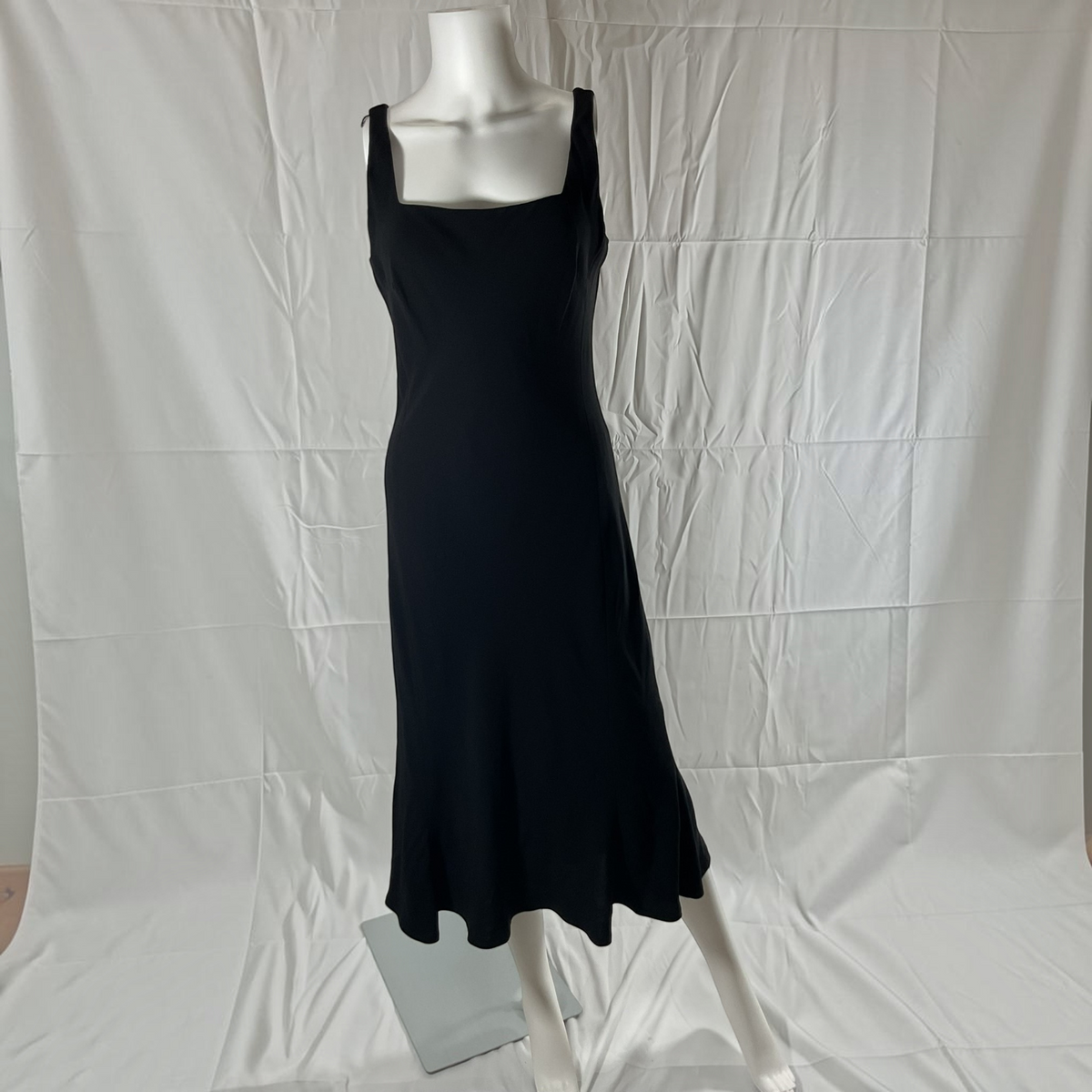 A black sleeveless midi dress with a square neckline and wide shoulder straps, displayed on a white mannequin against a wrinkled white fabric backdrop.