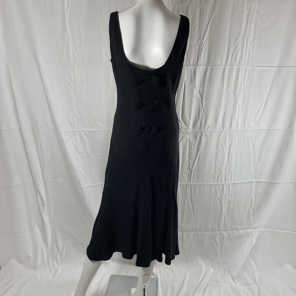A black sleeveless midi dress with a scoop neckline and three decorative bow details down the front, displayed on a white mannequin against a wrinkled white fabric backdrop.