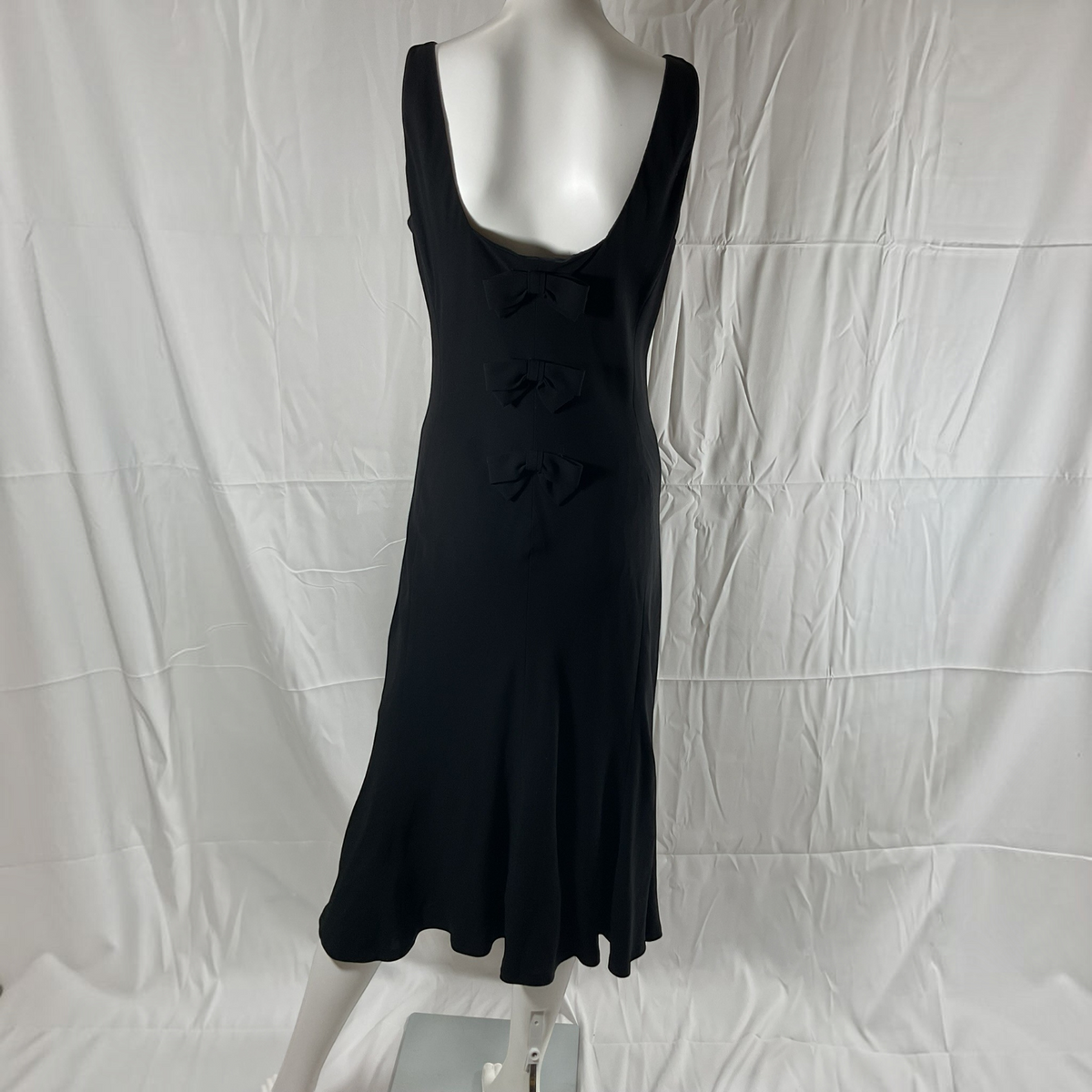A black sleeveless midi dress with a scoop neckline and three decorative bow details down the front, displayed on a white mannequin against a wrinkled white fabric backdrop.