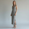 A woman modeling a sleeveless, floor-length striped knit dress with a high side slit, paired with white strappy high-heeled sandals, against a neutral light gray studio background.