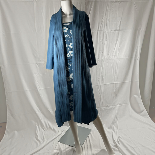 A blue pleated open-front cardigan or robe displayed on a mannequin, layered over a blue floral dress, against a white draped studio backdrop.