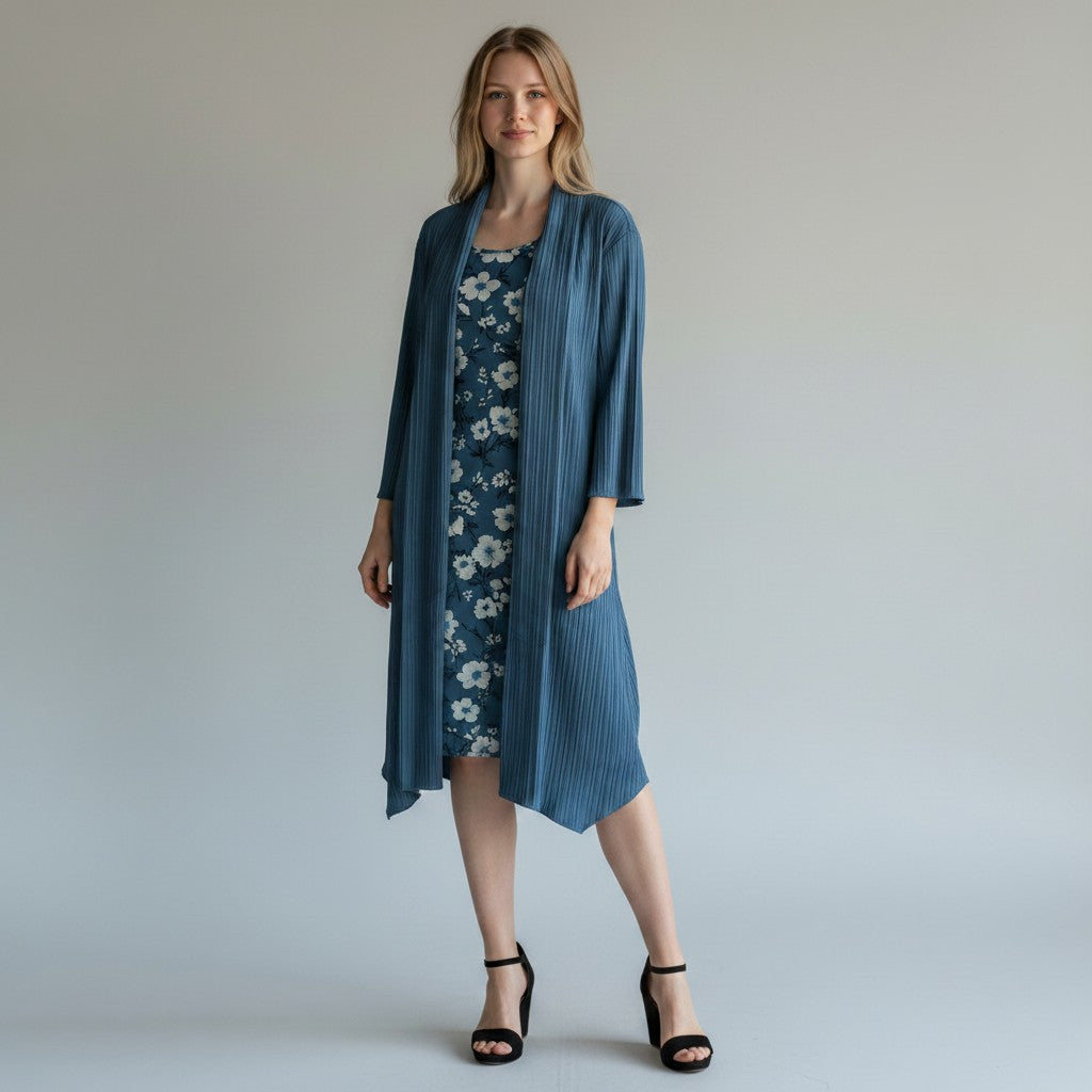 A woman modeling a slate blue pleated open-front cardigan over a navy floral midi dress, paired with black heeled sandals, against a light gray studio background.