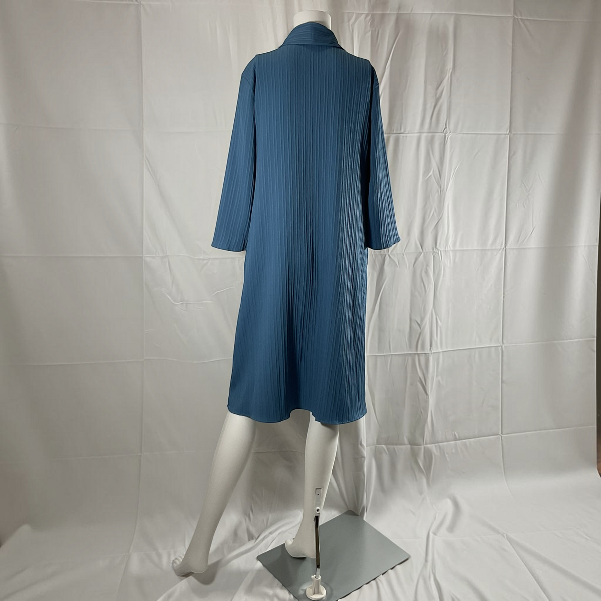 A blue pleated long-sleeve midi dress displayed on a white mannequin from the back, set against a wrinkled white fabric backdrop.