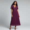 A woman wearing a short-sleeve midi dress in deep burgundy lace, paired with black strappy heels, against a light gray studio background.