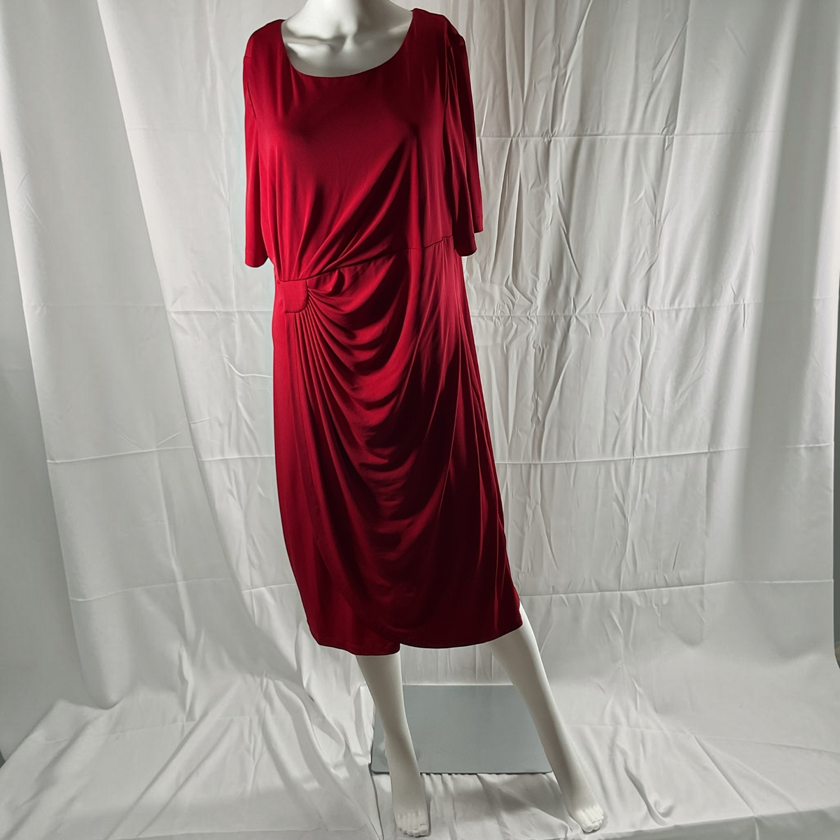 A red short-sleeve midi dress with a draped, gathered skirt detail, displayed on a white mannequin against a wrinkled white fabric backdrop.