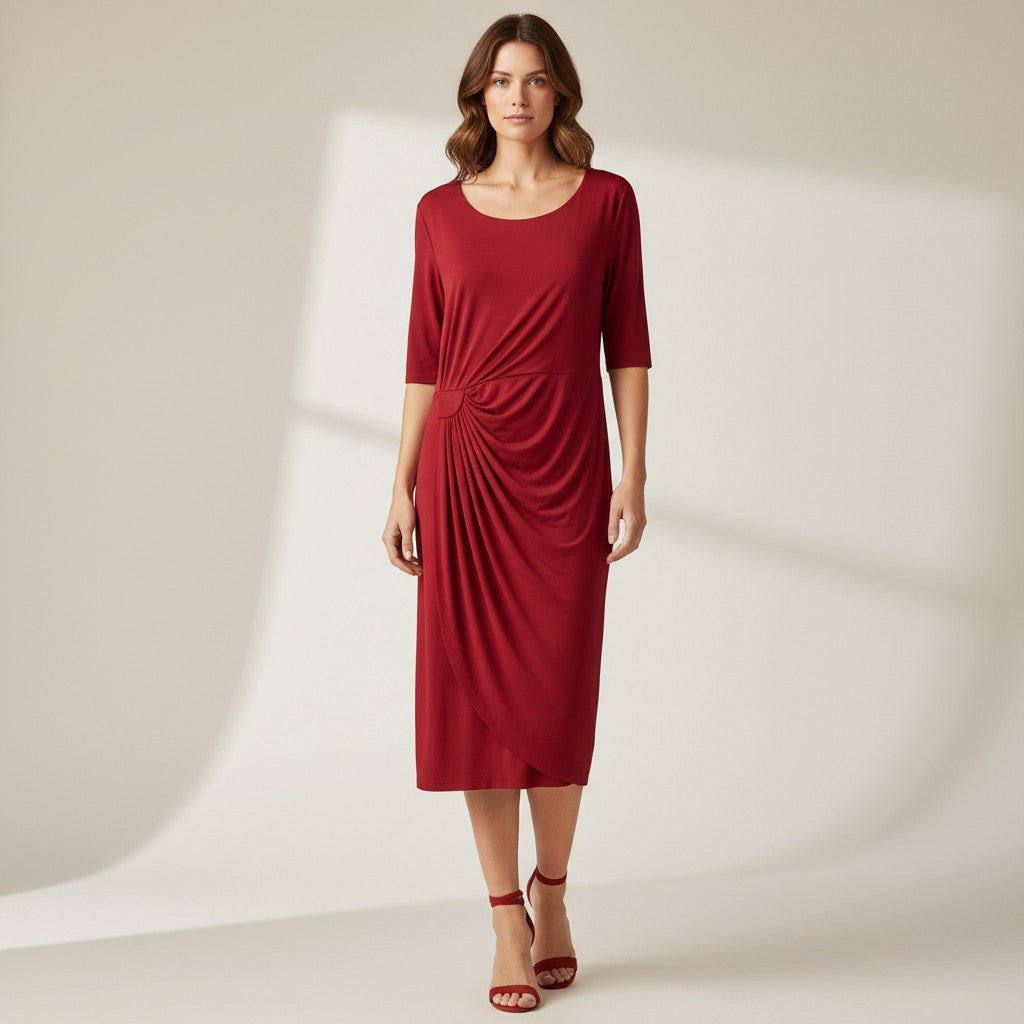 A woman modeling a red midi dress with elbow-length sleeves, a scoop neckline, and a gathered draping detail at the front, paired with red heeled sandals against a warm off-white studio background.