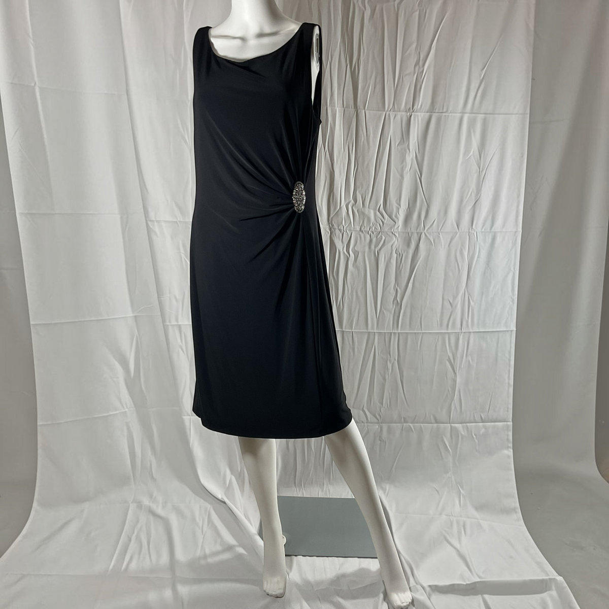 A sleeveless black midi dress with a gathered twist detail at the waist and a decorative silver-toned brooch, displayed on a white mannequin against a wrinkled white studio backdrop.