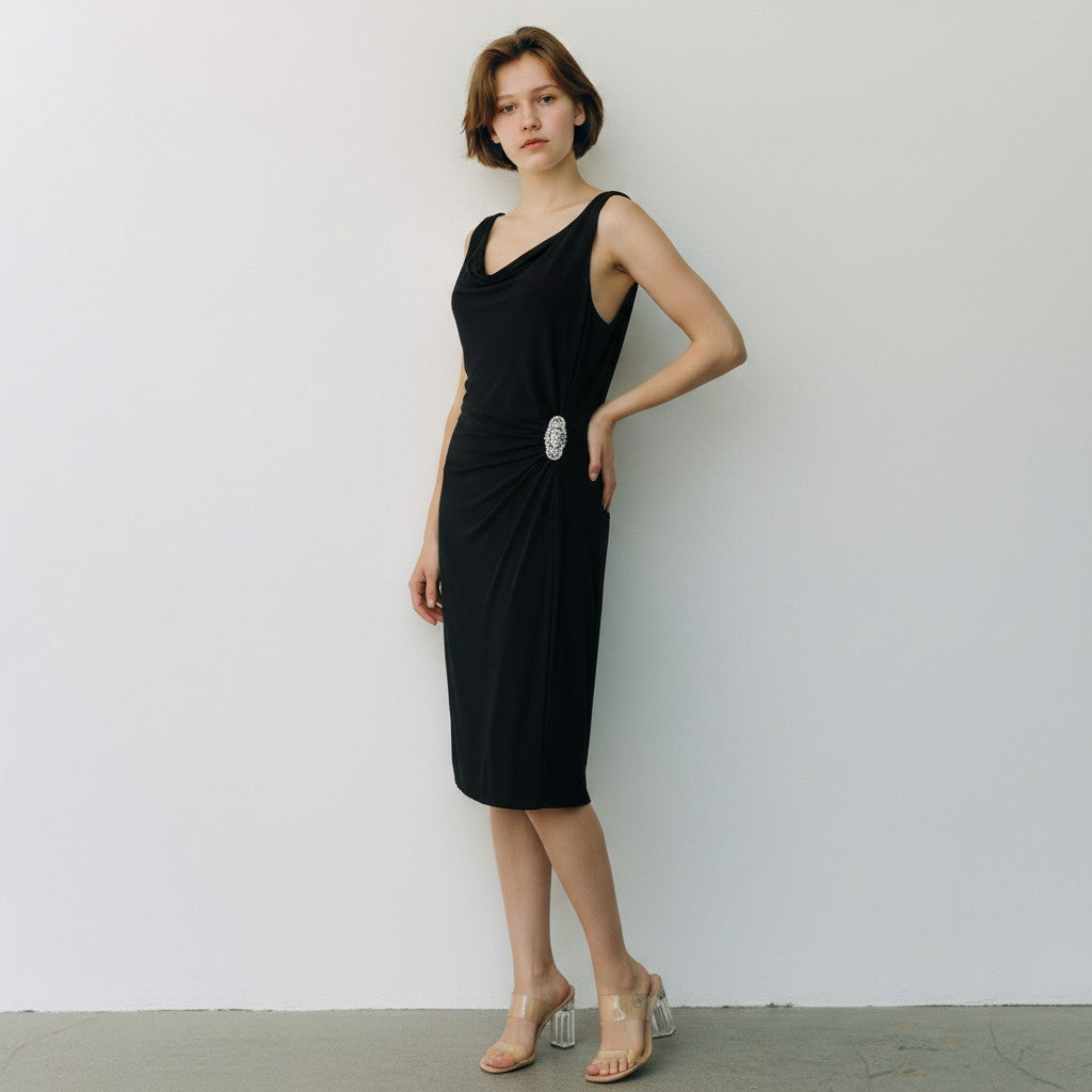 A woman modeling a sleeveless black midi dress with a draped neckline, gathered side detail, and a large silver oval brooch, standing against a plain light gray wall.