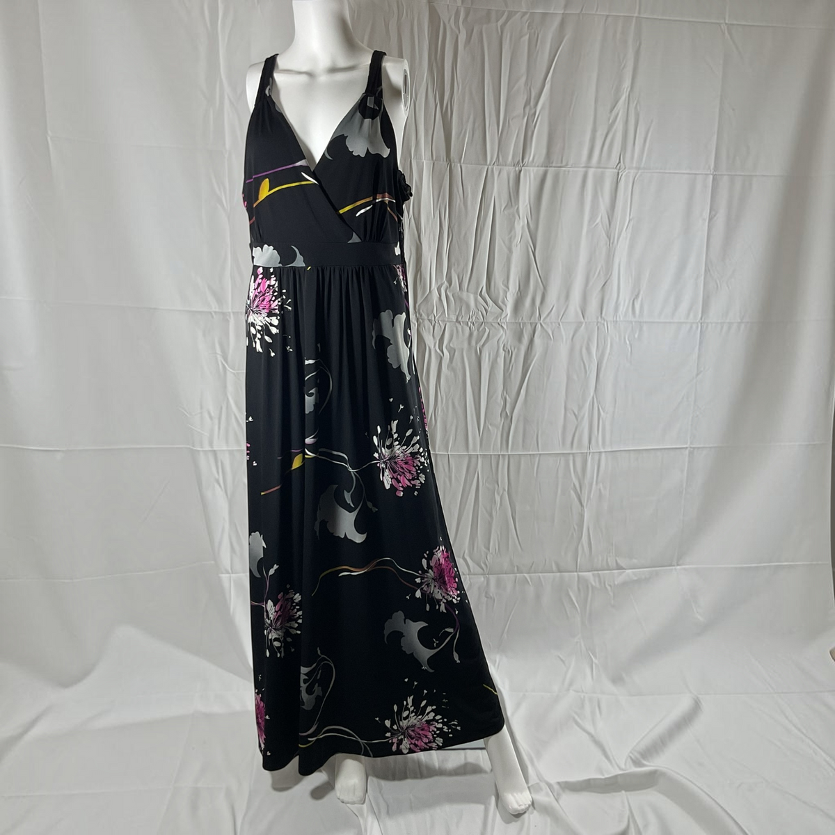 A sleeveless maxi dress displayed on a white mannequin against a wrinkled white fabric backdrop. The dress has a black base with a floral and abstract print in gray, pink, and yellow.