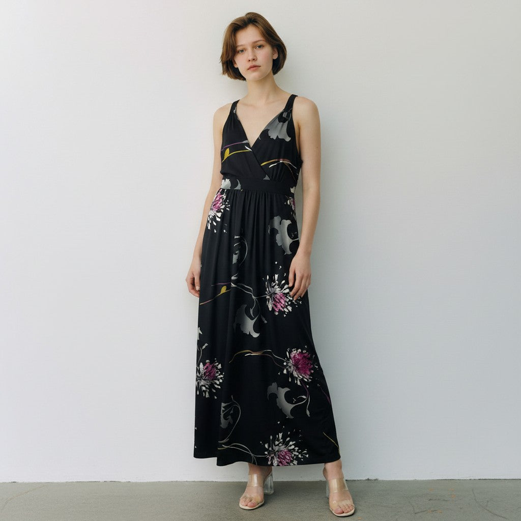 A woman stands against a plain light gray wall wearing a sleeveless black maxi dress with a floral and abstract print in pink, gray, and gold, paired with clear high-heel sandals.